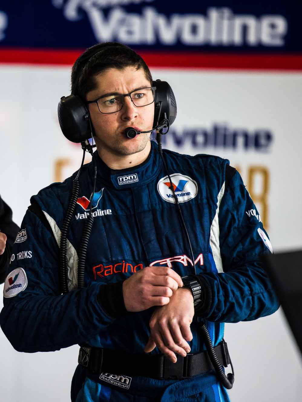 Stefan has short brown hair and wears thin black framed glasses. He is wearing a navy jumpsuit with sponsor logos on it. He is wearing a headset with microphone and is looking at his watch.