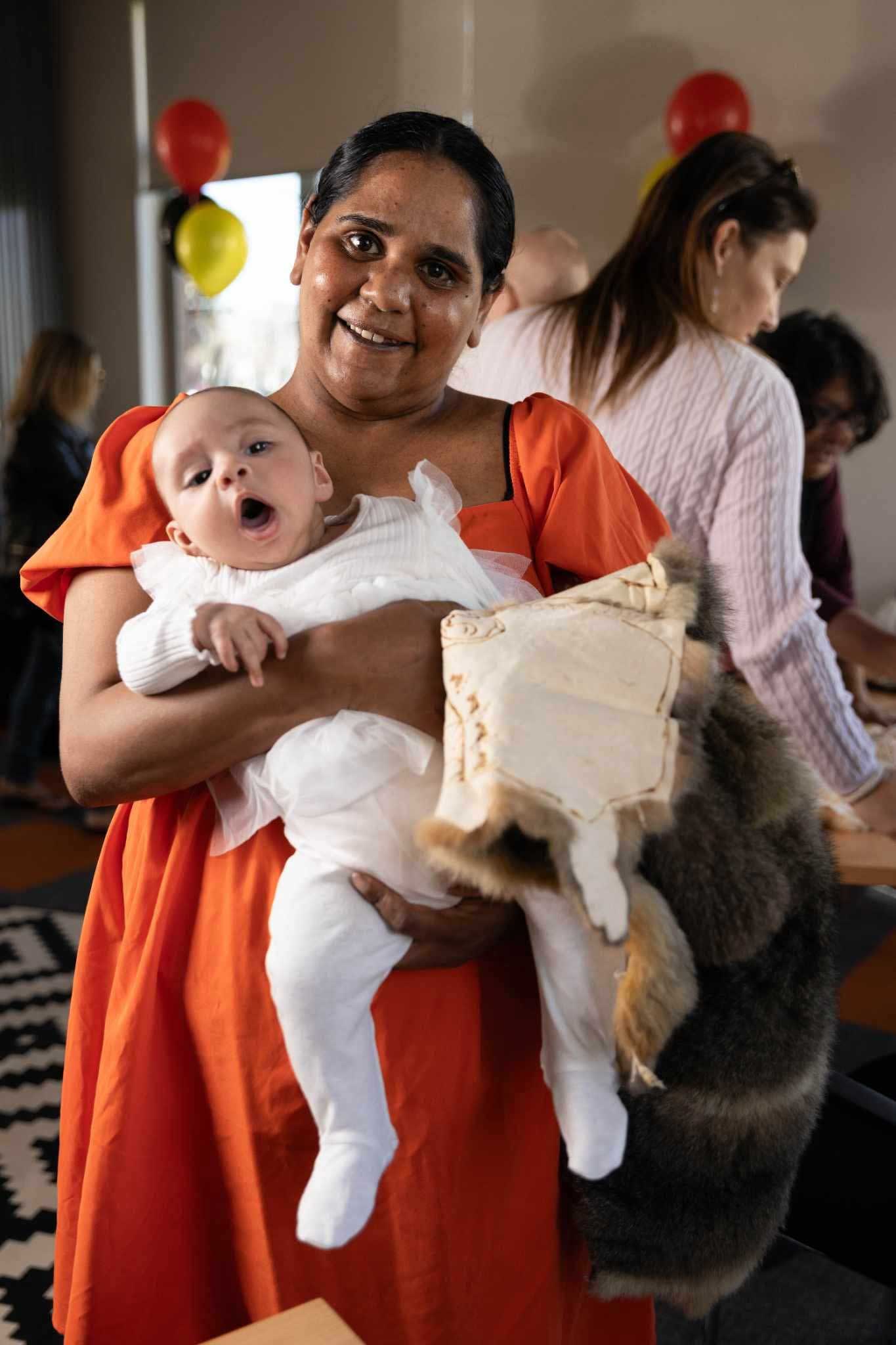 Eugenie is wearing an orange summer dress and is holding her baby daughter who is wearing a white onesie.