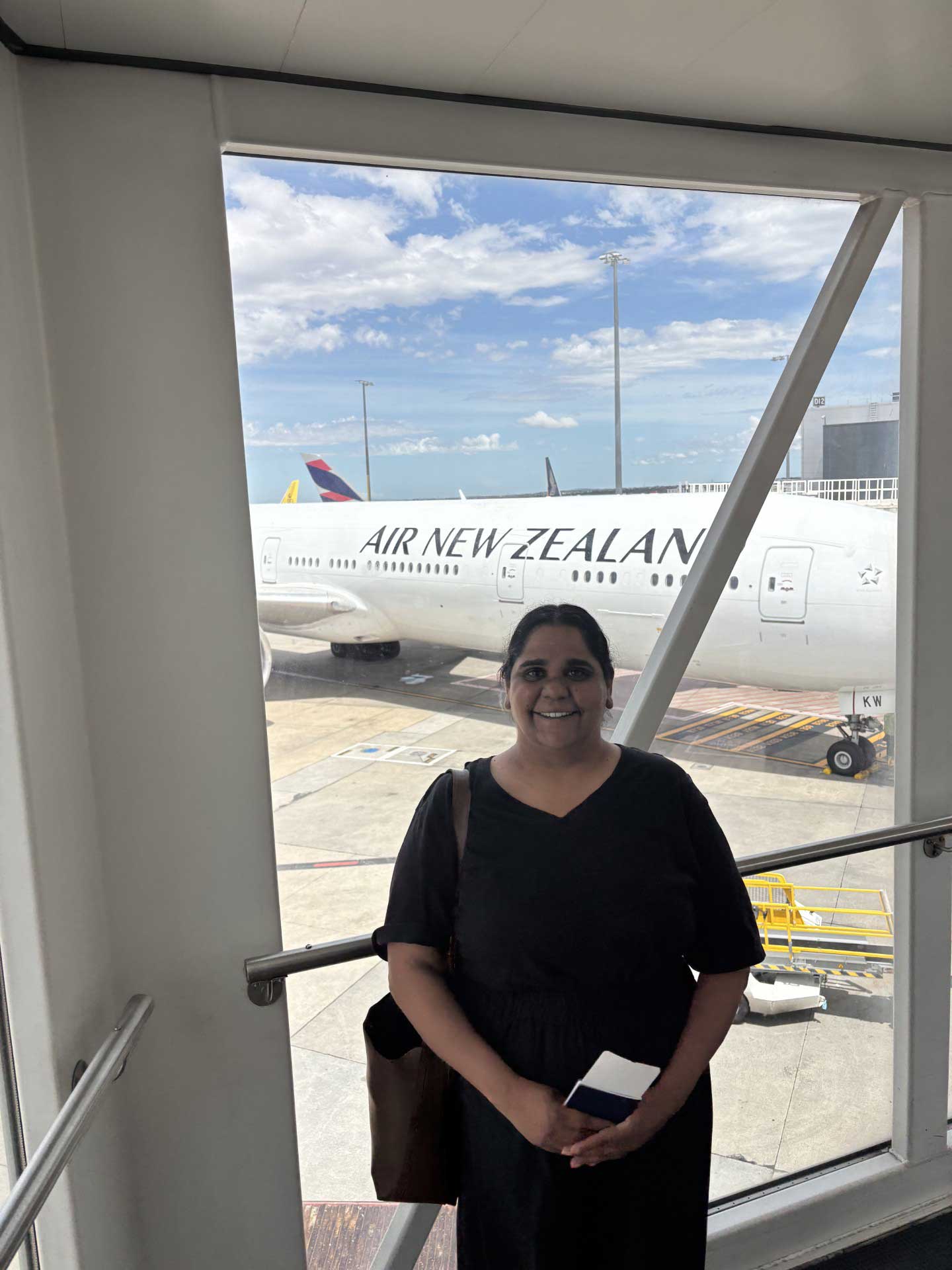 Eunice is standing in front of a window with an Air New Zealand aircraft in the background. She is wearing a black dress and carrying a tote bag.