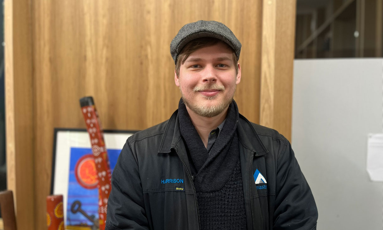 Harrison has a short beard and is wearing all black. He is wearing a black flat cap.