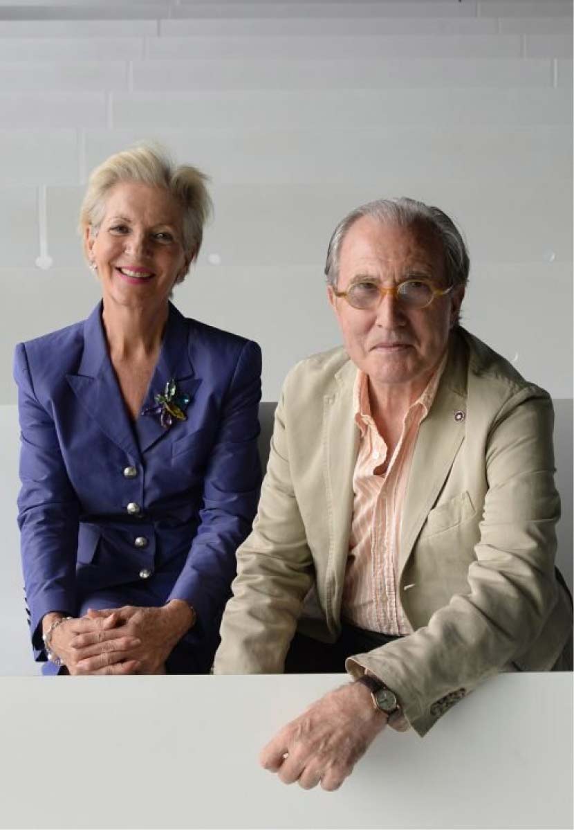 A phot of Annmarie on the left and Arturo on the right. Annmarie has short blonde hair and is wearing a fitted purple linen suit with a broach on the lapel. Arturo is wearing tan coloured spectacles and a beige linen suit.
