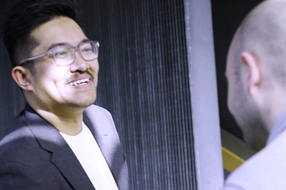 Two people standing and talking in an indoor, modern environment with dark walls and lighting, wearing business casual clothing.