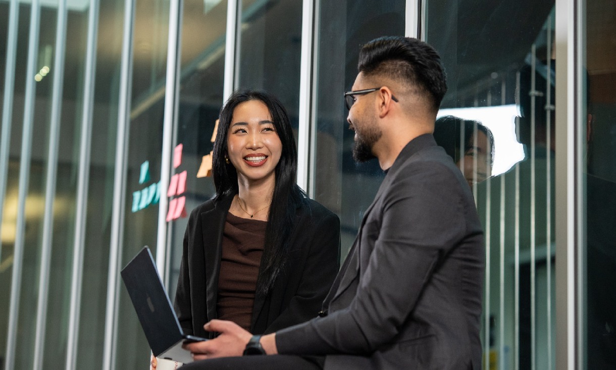 Two people in business attire sit and talk in front of tall glass windows, one holding a laptop and phone.