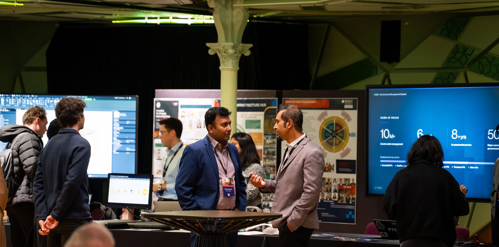 Two people talking at an industry event with posters and large screens behind them. 