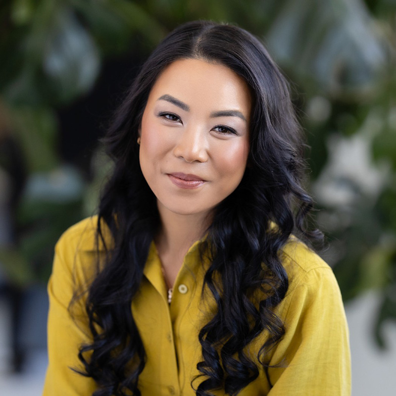 Photograph of Alyna Pham. She smiles directly at the camera and. She has long, black and wavy black hair and wears a yellow shirt.