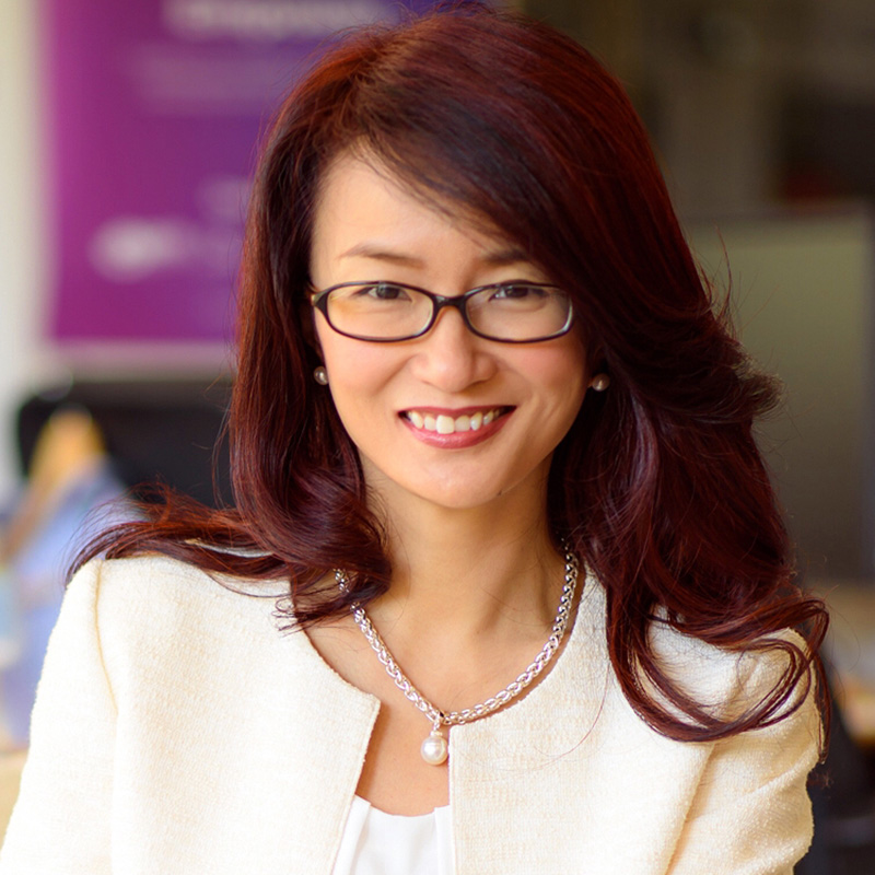 Photograph of Dr Sandy. She smiles directly at the camera. She has long, dark red hair and is wearing glasses. She is also wearing a white jacket.