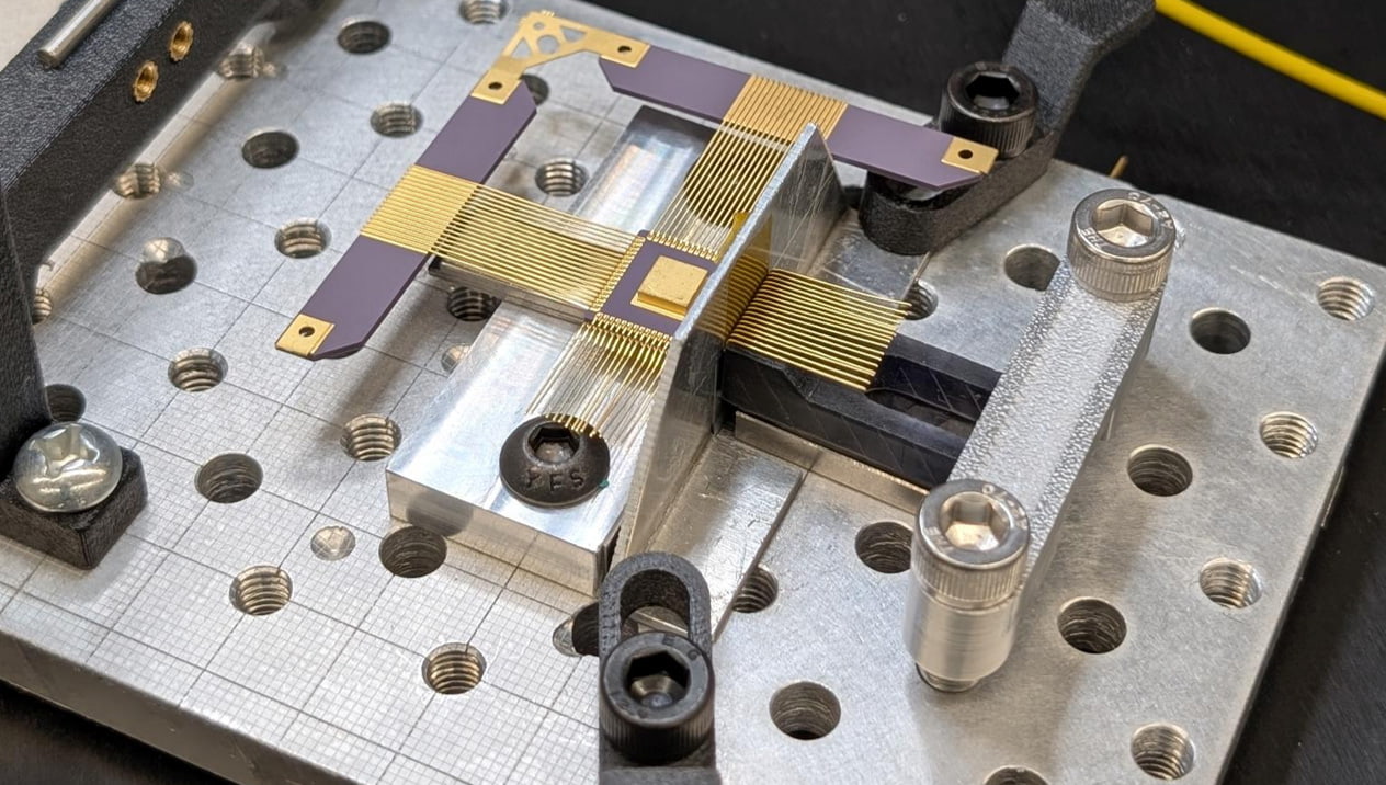 Figure 4 shows a close-up, top-down view of a highly precise electronics assembly.  Here is a breakdown of what is visible in the image:  The Fixture and Breadboard  Optical Breadboard: The entire assembly is mounted onto a silver, metallic grid plate (an optical breadboard or fixture plate) featuring a pattern of threaded screw holes.  Clamps: Several black and silver mechanical clamps, held down by hex screws, are securing the component setup firmly in place. Central Element: At the center of the fixture rests a delicate micro-electronic component called an FPGA. It features a tiny, square gold pad in the middle.  Fine Wire Arrays: Radiating out from the central area are arrays of extremely fine, parallel gold wires or traces that bridge across a small gap.  Supporting Wings: These delicate wire arrays are connected to purple, wing-like structural tabs with gold contact points at their tips.
