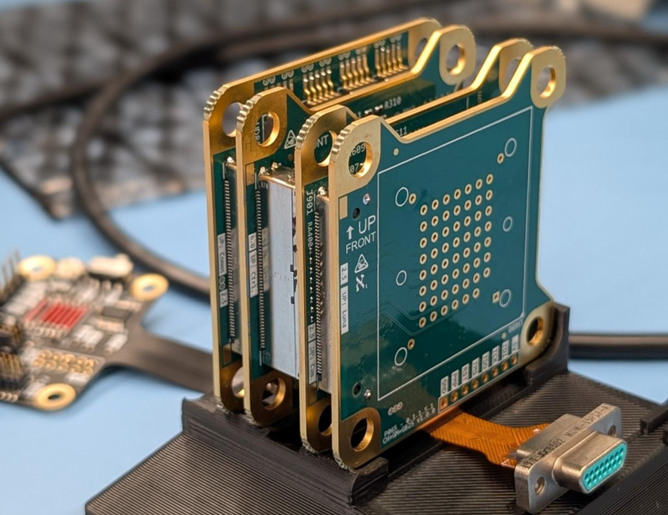 Figure 6  shows a close-up view of a hardware electronics setup on a light blue work surface. Here are the key details visible in the image:  The Main Assembly: In the center, several small, green printed circuit boards (PCBs) with gold-plated edges and mounting holes are stacked vertically next to each other. They are held upright in a black, 3D-printed plastic dock or stand.  Markings and Components: The outermost visible PCB has grid-like pin holes. A ribbon cable extends from the bottom of the board assembly, ending in a metal D-sub connector.  Background Elements: Another smaller, flat circuit board with attached wiring is visible to the left. The background is softly blurred but shows lab or workshop equipment, including what appears to be a computer keyboard, cables, and electronic testing gear.