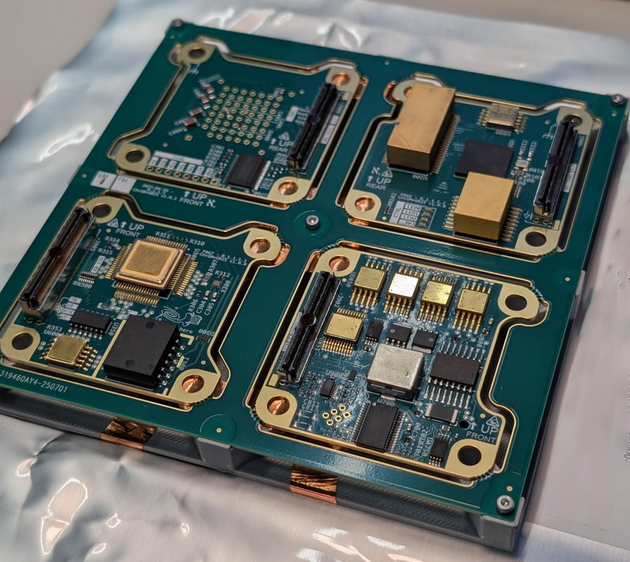 Figure 5 shows a detailed, top-down view of a complex electronic assembly consisting of a multi-board printed circuit board (PCB) panel resting on an anti-static surface. Here is a breakdown of what is visible in the image: Board Layout and Structure  Four-Quadrant Design: The main green PCB is a large panel or motherboard divided into four distinct quadrants, each holding a smaller, integrated circuit board module.  Gold-Plated Borders: Each of the four sub-board areas is framed by thick, gold-plated routing lines and features rounded corner tabs with circular mounting holes, similar to the individual boards seen in image.png.  Structural Framing: The entire green board assembly is secured within a gray, precision-machined metal frame or carrier, with copper-colored grounding tabs visible along the bottom edge. Electronic Components Each quadrant contains a unique arrangement of high-density surface-mount components:  Connectors: Long, black, multi-pin board-to-board connectors are mounted on several of the modules.  Integrated Circuits (ICs): There are various black microchips, resistors, capacitors, and small surface-mount devices scattered across the boards.  Specialized Components: Several metallic, gold-colored blocks or shielded packages (potentially sensors or specialized processors) are prominent on three of the four modules.