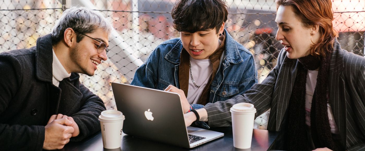 hree students sitting at a table outside, all of them looking at a laptop screen, takeaway coffee cups waiting to be sipped.