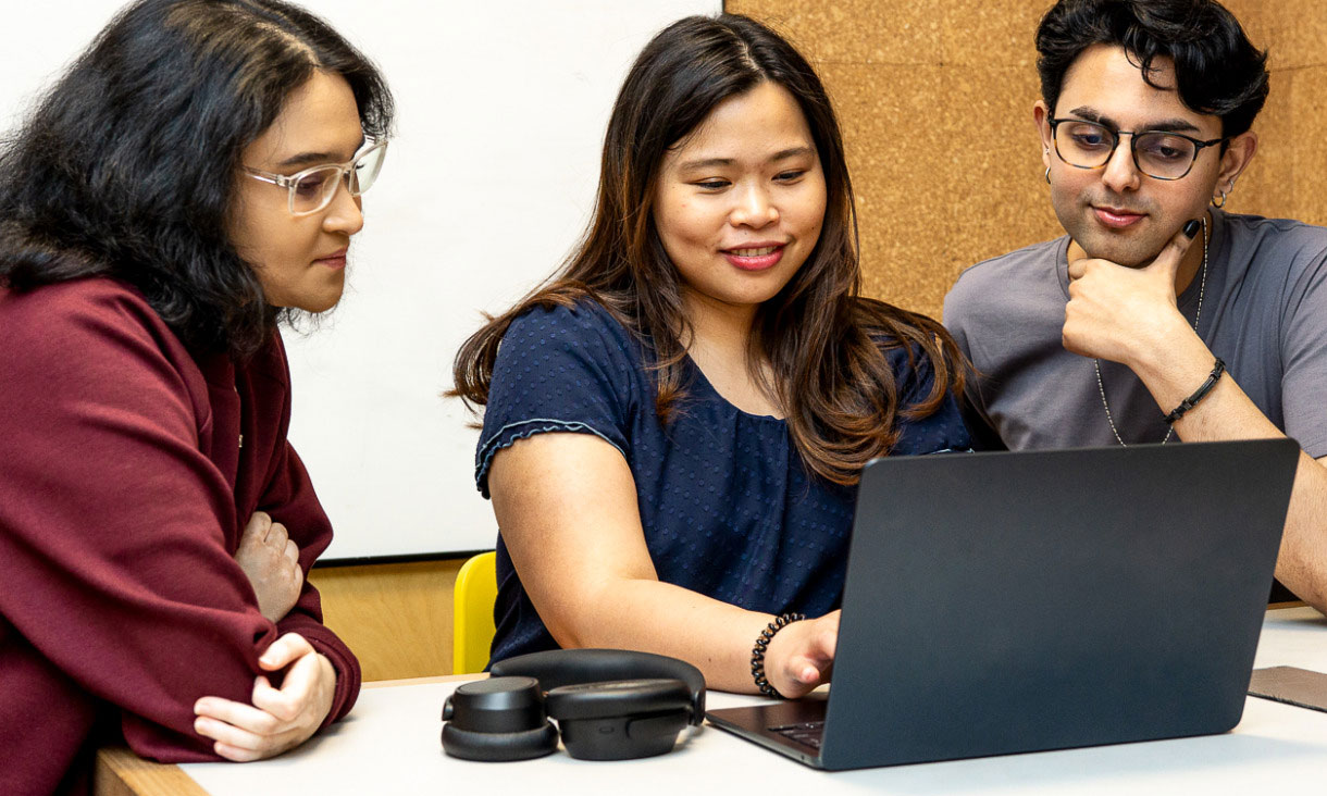 Three students collaborate around a laptop in a Carlton group study room.