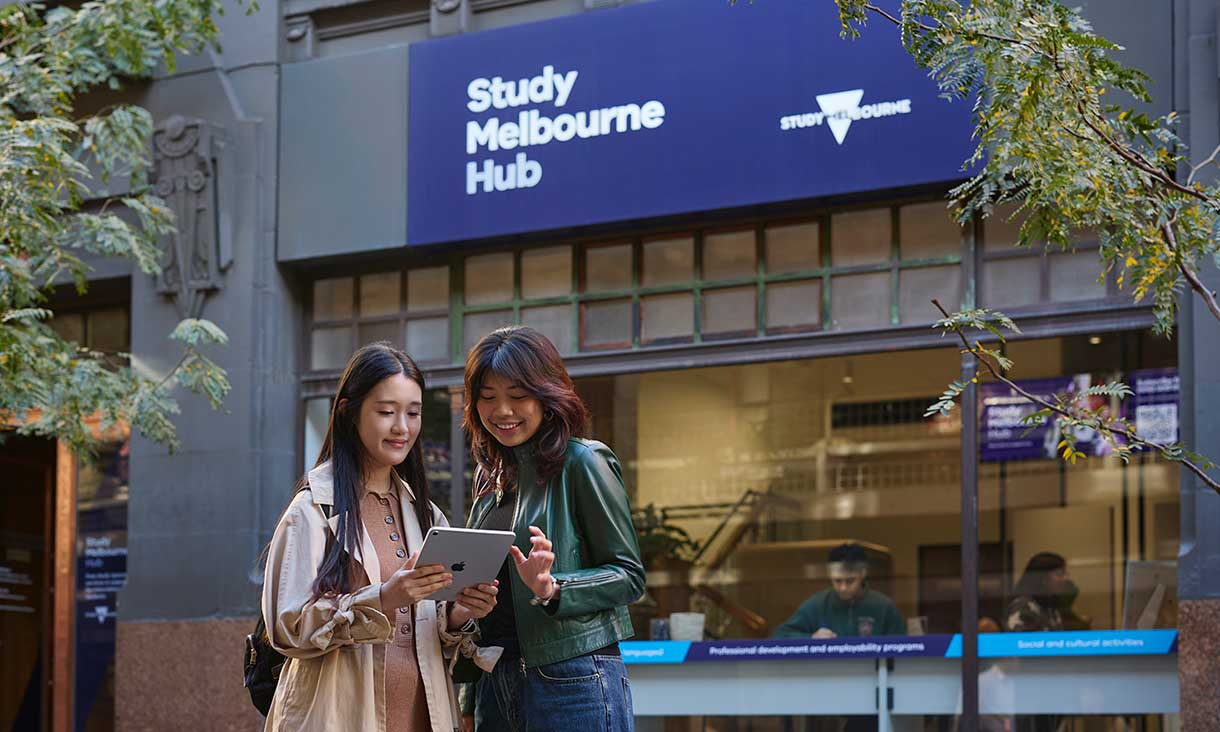 Two students standing outside the Study Melbourne Hub, looking at a tablet together and smiling, with the hub’s blue sign visible above the entrance.