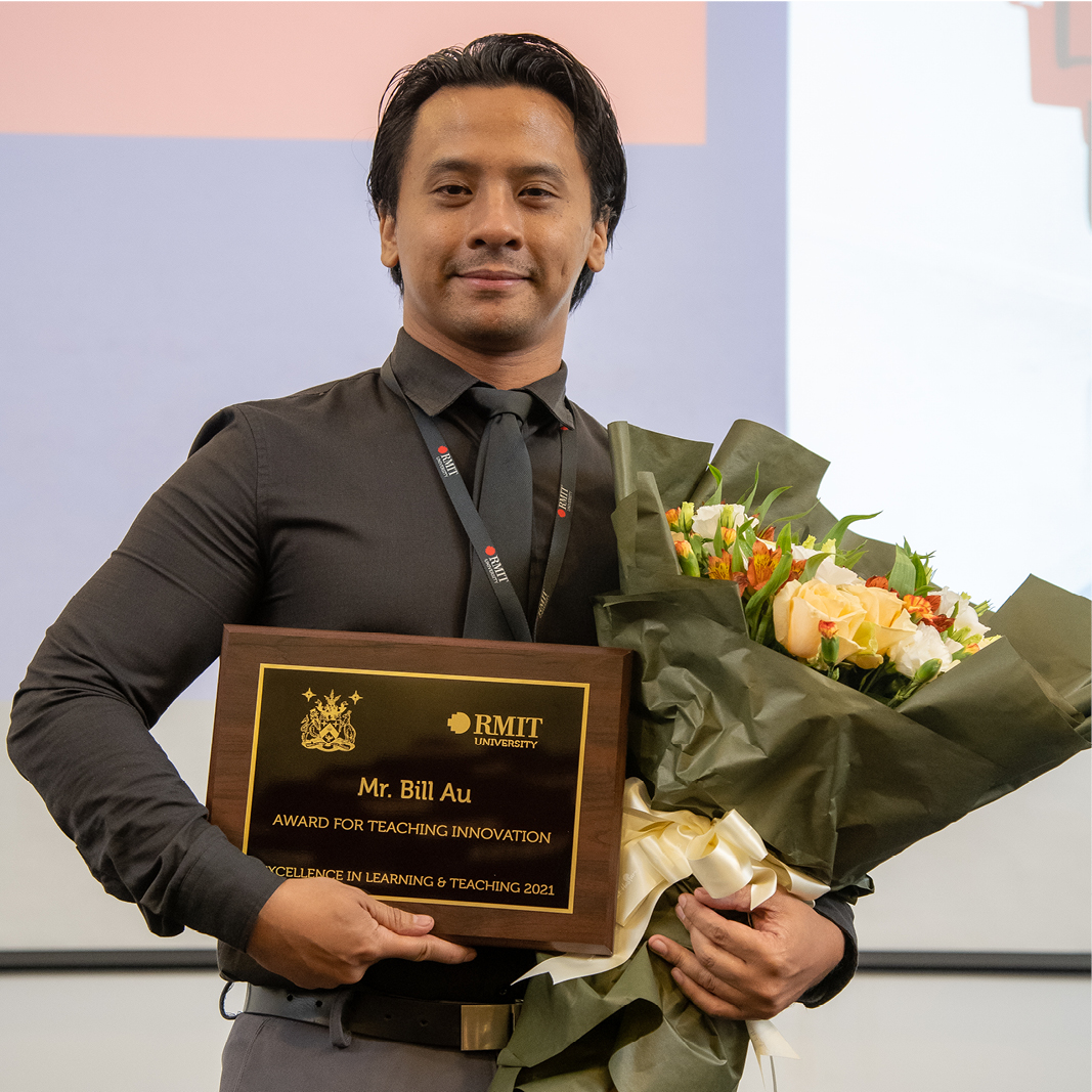 A man is smiling at the camera and holding a plaque and bouquet of flowers. Plaque reads Mr BIll Au, Award for Teaching innovation 