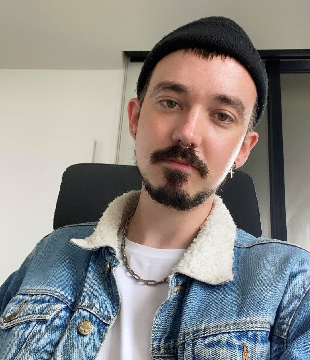 A man is looking directly at the camera. He is wearing a beanie and has a nose ring and facial hair.