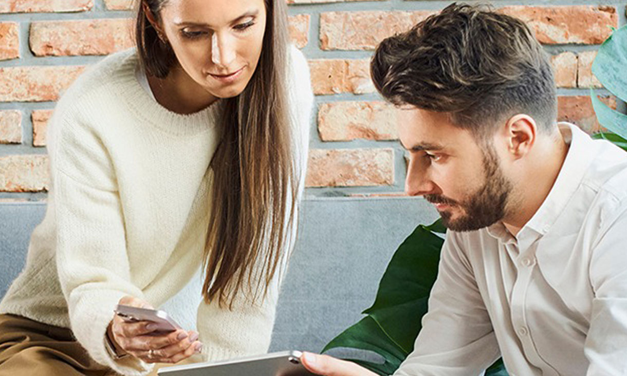 A woman shows a man her phone in an office while he holds a tablet. 