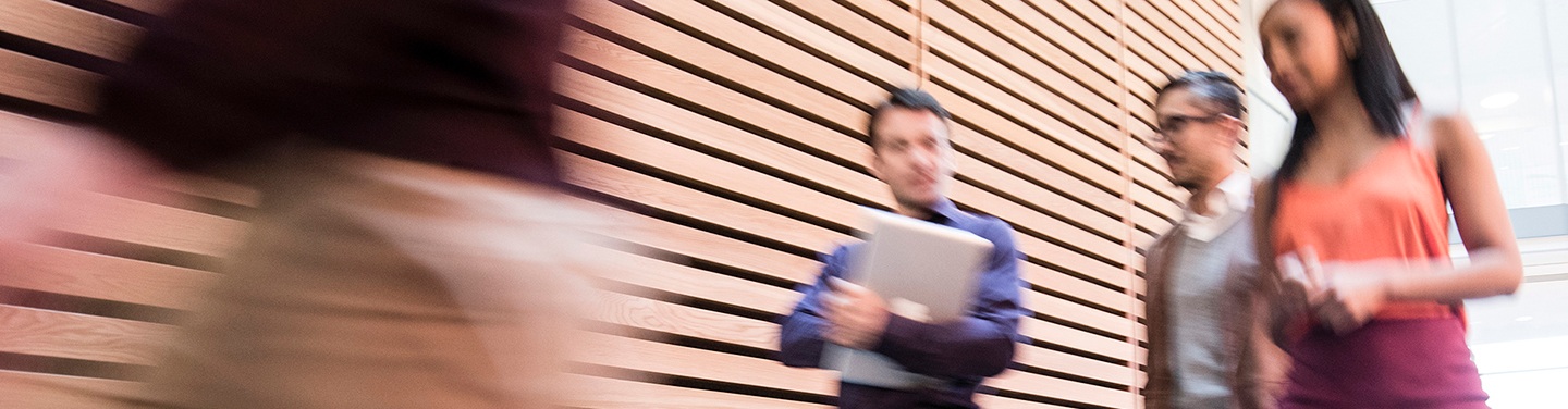 A group of people walk past a wall with wooden slats in an office.