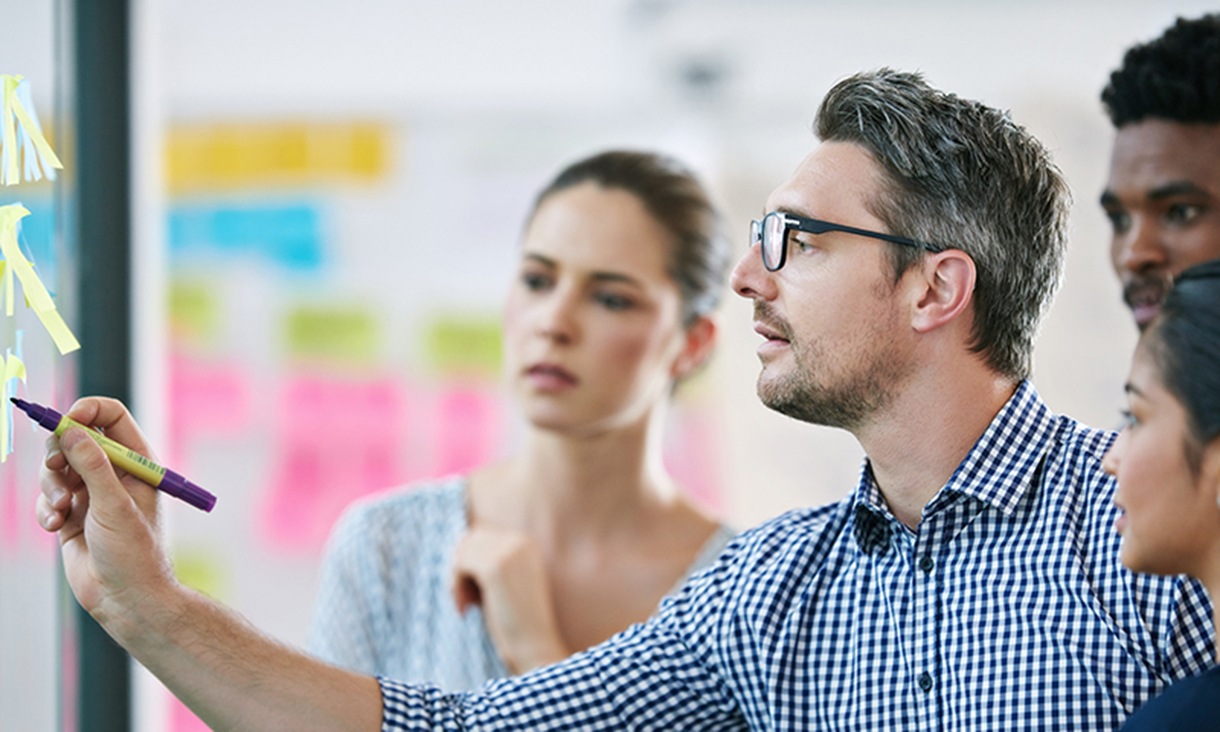 A man makes a note on a wall of sticky notes while being watched by others during a meeting.