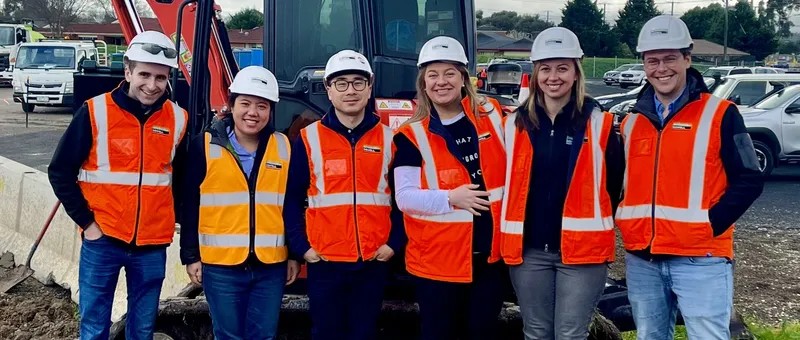 A group of people wearing hard hats and high-vis vests standing in front of a digger on a construction site.