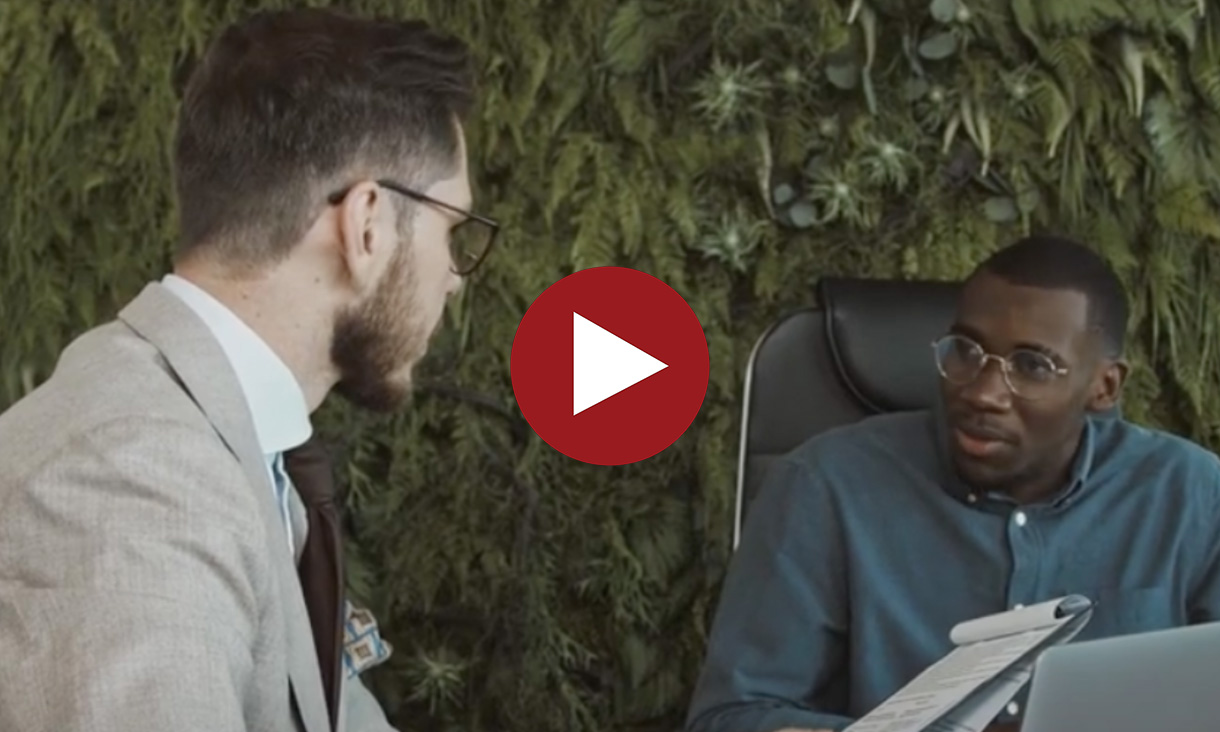 Two men having a discussion in a modern office setting, one holding papers, with a red play button overlaid in the centre.