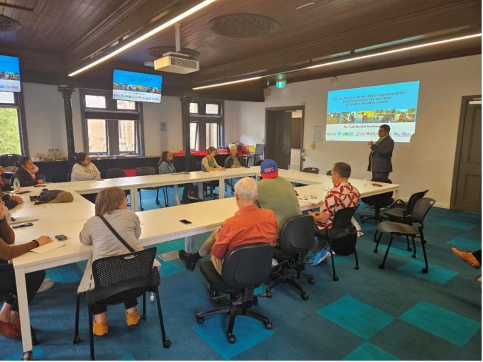 A man standing before a screen at the front of a room presenting to a medium sized group who around a large rectangular table.