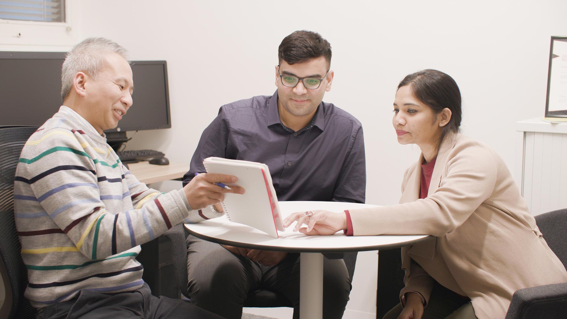 Three people sit around a table looking at what is written on a notebook