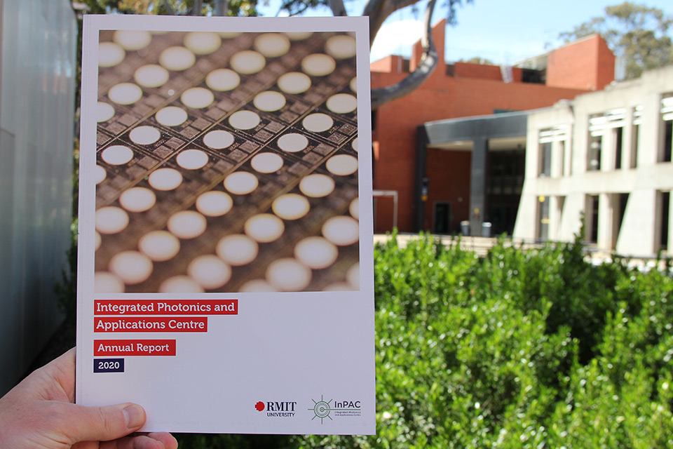 A hand holding up a report with the words 'Integrated Photonics and Applications Centre Annual Report 2020'