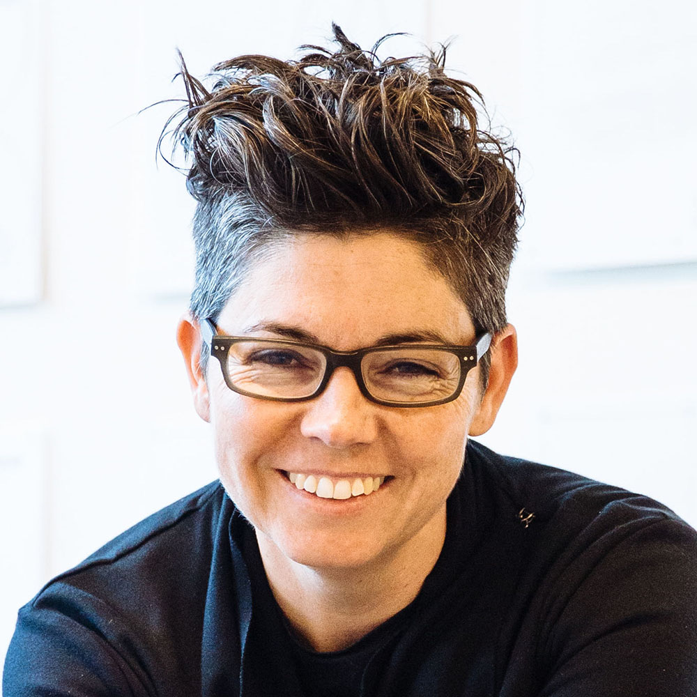 Photograph of Naomi Stead. She is smiling directly at the camera. She wears glasses with black frames and wears a black shirt. She has short hair that is styled to be swept up and back away from her face.