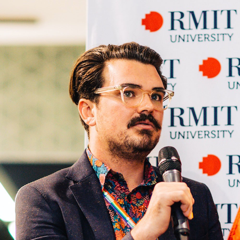 Photograph of Patrick Kelly. He is looking away from the camera and his holding in microphone in his hand. He wears a pair of glasses with clear frames and his dark hair is swept neatly away from his face. Behind him is a backdrop with the RMIT logo.