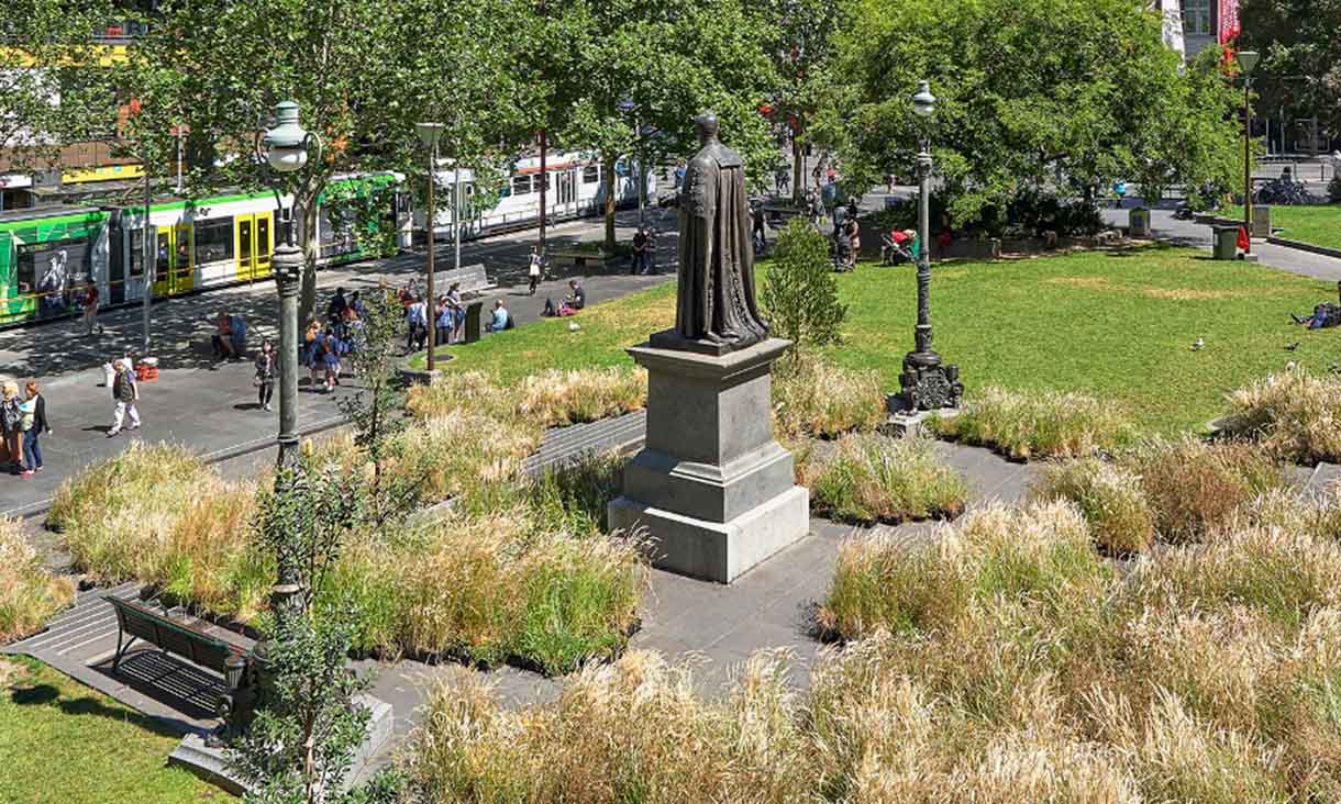 Photograph of the front of the Melbourne State Library. In the centre of the photograph is the Redmond Barry, which is facing away from us. The concrete path is covered by grassy bushes.