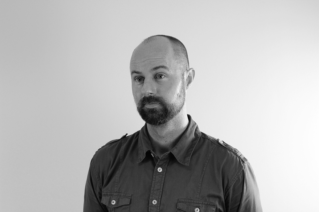 Black and white headshot of Daniel Scow. He has a short beard and wears a button up shirt.