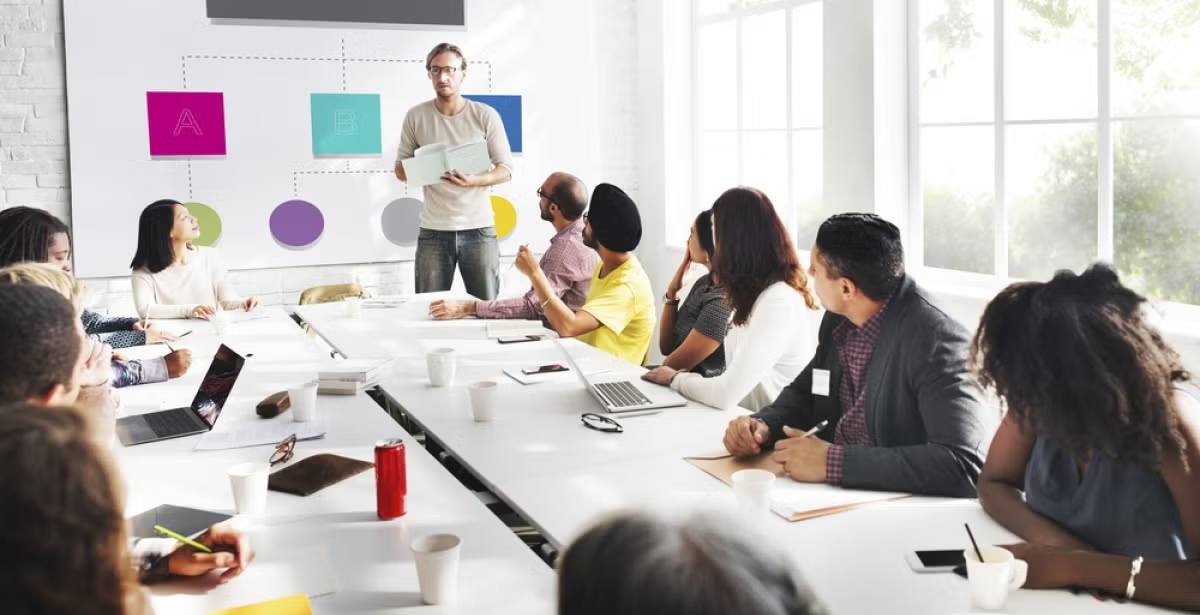 12 people sitting around a table with their laptops and notebooks watching while one stands to speak. 