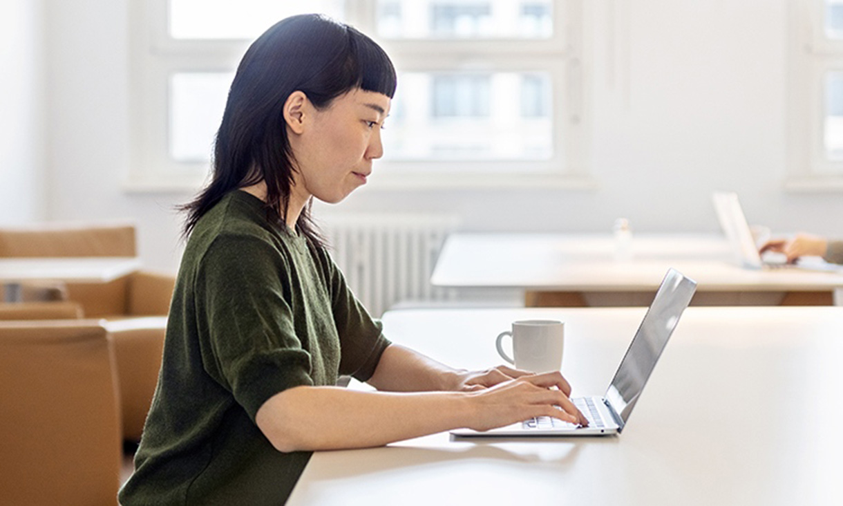 A woman on her laptop in the foreground with someone on their laptop in the background.