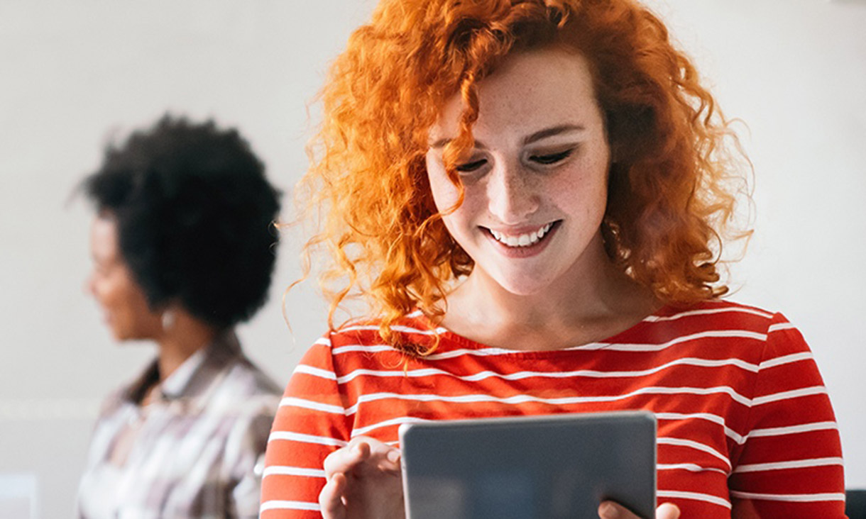 A woman on a tablet in the foreground with a colleague in the background.