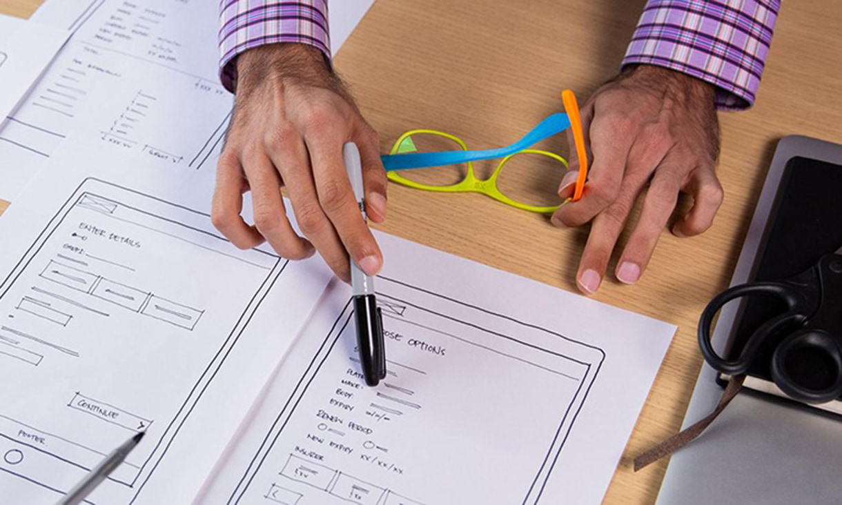 Hands holding glasses and a pen pointing at a UX smart phone plan.