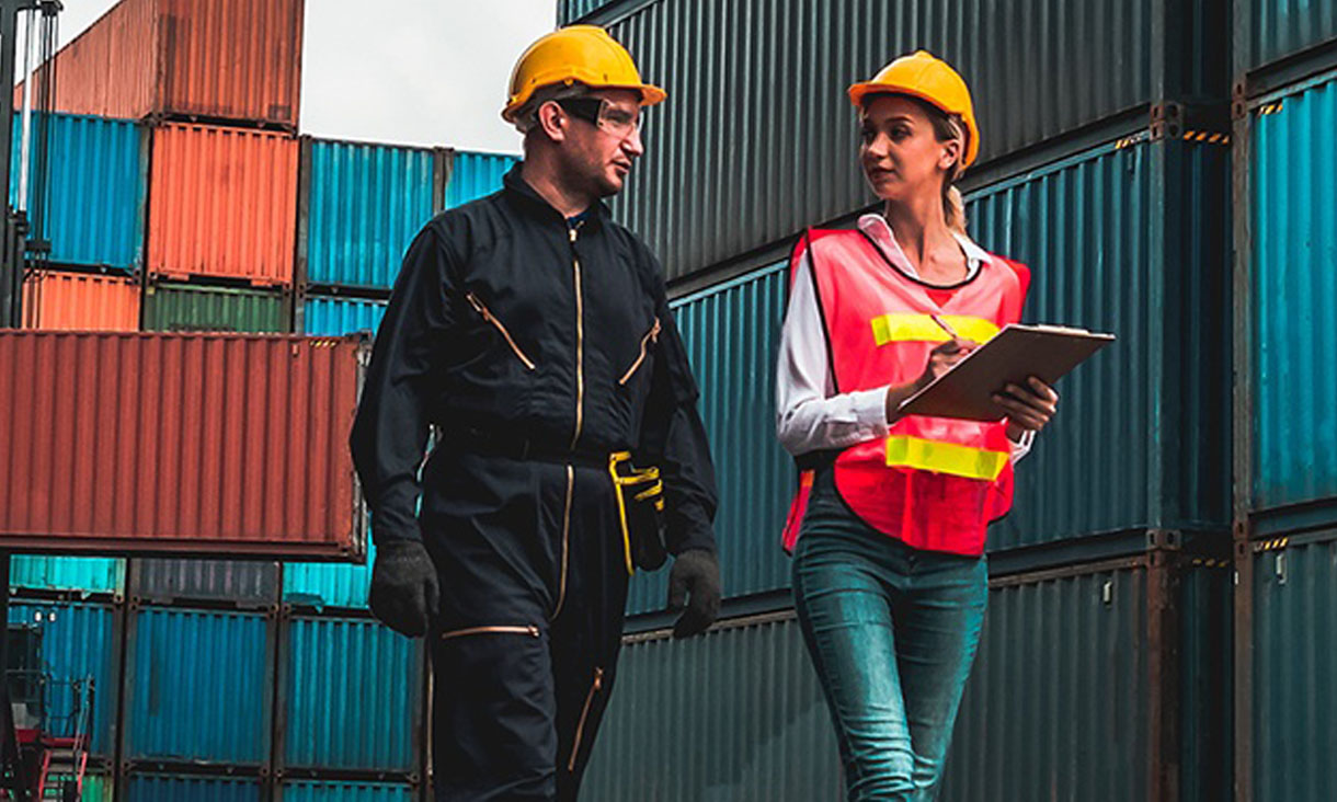A man and a women walk through a shipping container lot. They both have on hard hats and the woman has a clipboard. 