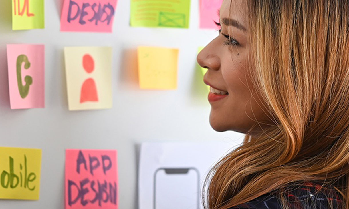 A woman stands at a wall that has a mixture of post it notes and iphone app deisgns stuck up. 
