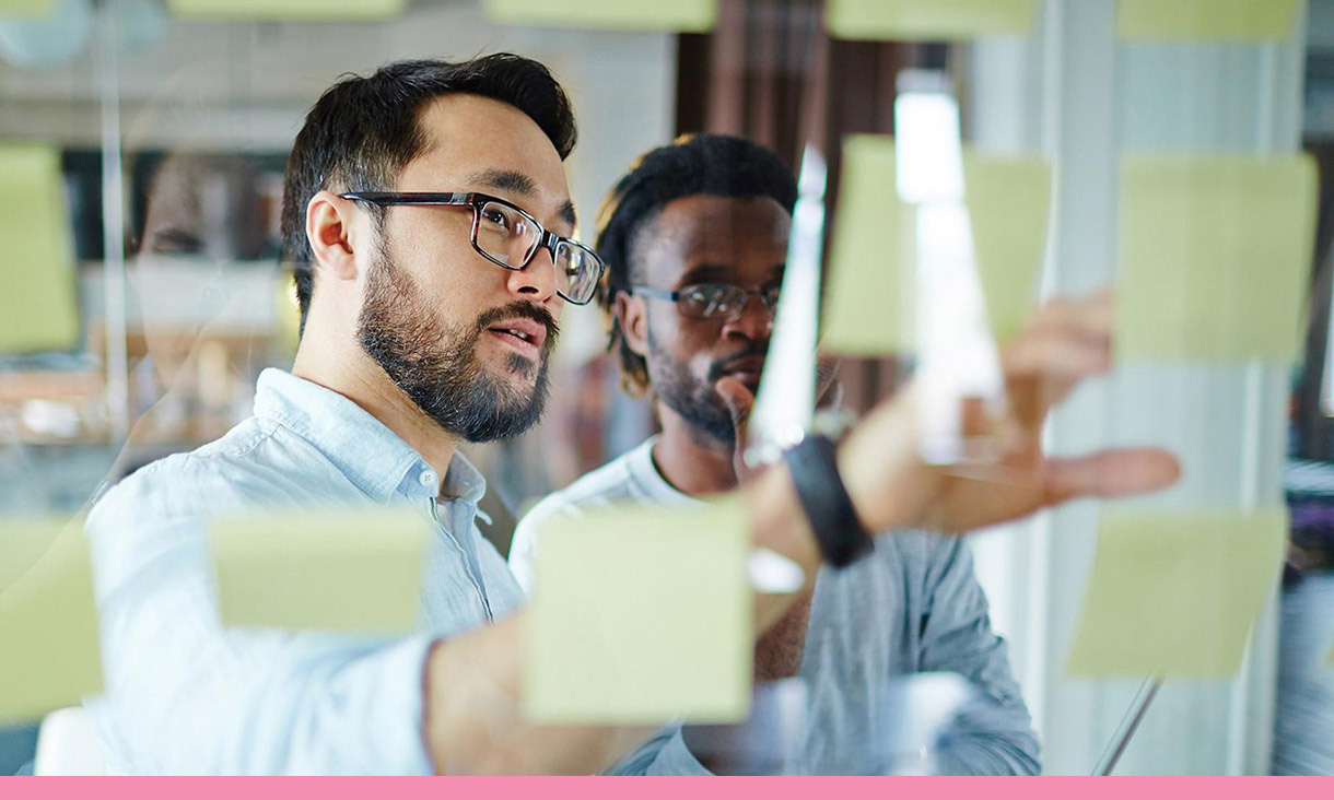 2 coworkers examine a board of sticky notes.