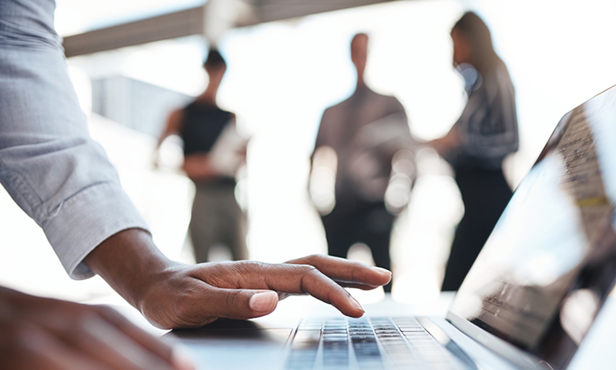 A person types on their laptop in the foreground while three people stand around in the background.