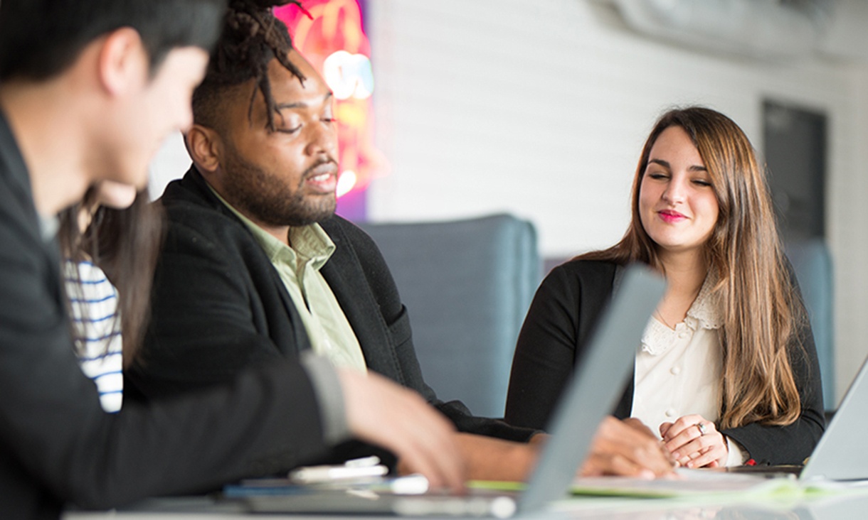 Four people sit at a shared desk during a meeting.