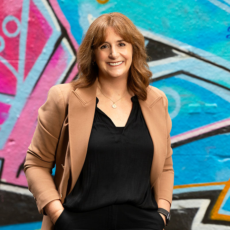 Photo of smiling woman dress in a black top and a tan jacket, standing in front of a wall mural.