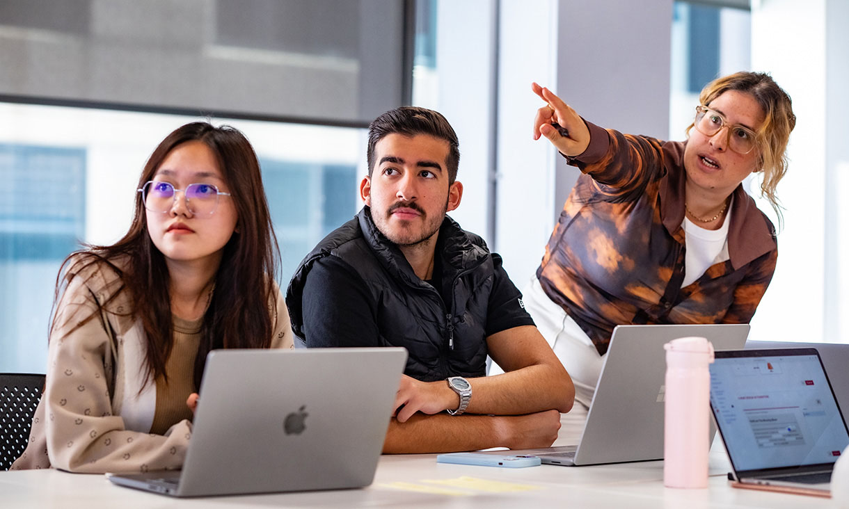 Three students in a English classroom, two are seated and one is pointing towards.