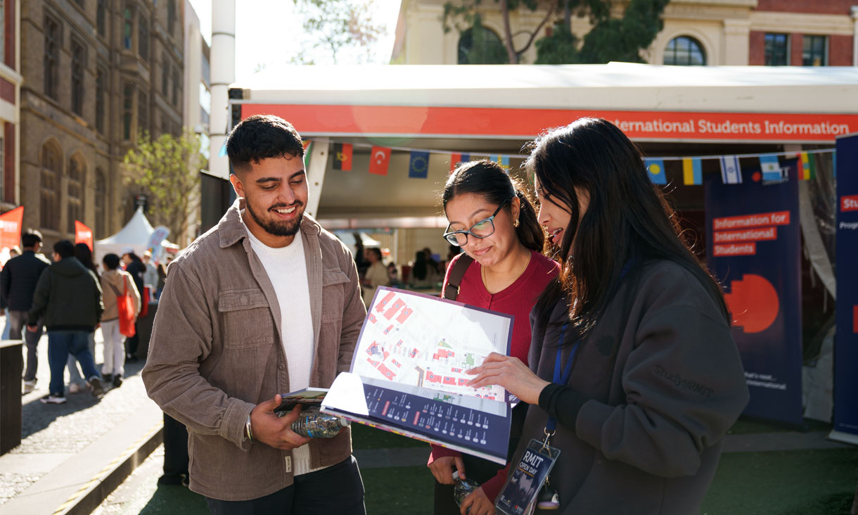 Three people at an outdoor RMIT International Students Information booth, looking at a campus map and engaging in conversation, with informational banners and flags in the background.