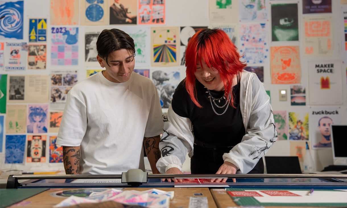 Two arts students working in a studio with prints on a wall behind them