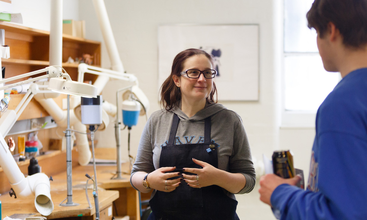 A student wearing overalls talks to another student, standing in front of a pipe model. 