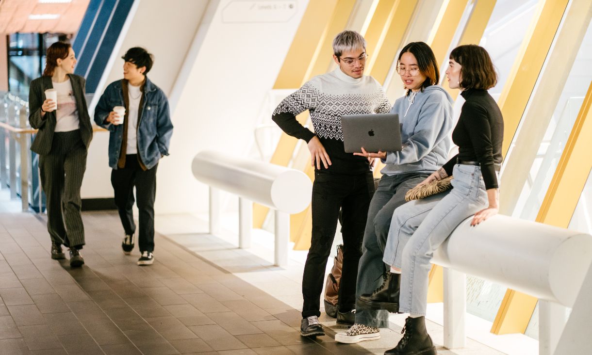 A group of students on campus looking at a laptop. Some people with takeaway coffee cups chatting in background