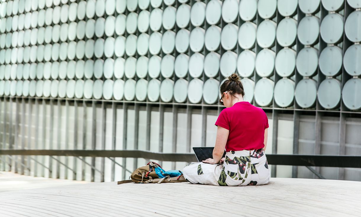 A person sitting on a wooden deck using a laptop beside a backpack, with a modern building facade featuring rows of circular panels in the background.