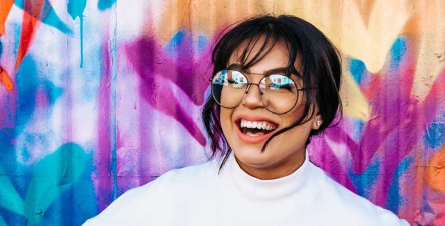 A student wearing glasses smiles. They're standing in front of a colourful wall spray painted with purple and blue.