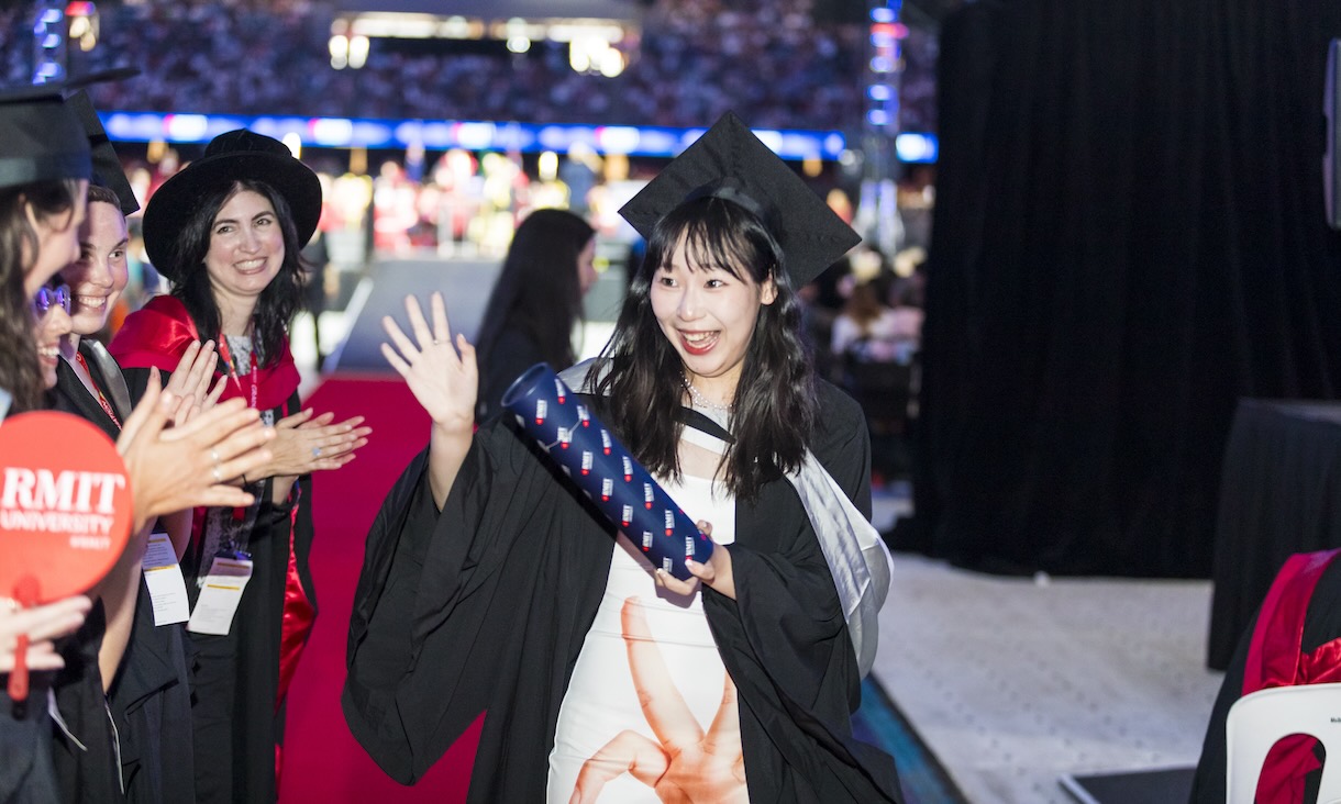 Female graduate looking at camera and holding a flag in front of people and a doughnut van on Bowen Street