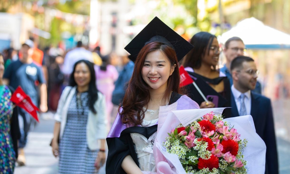 Female graduate stands among a crowd smiling, holding flowers