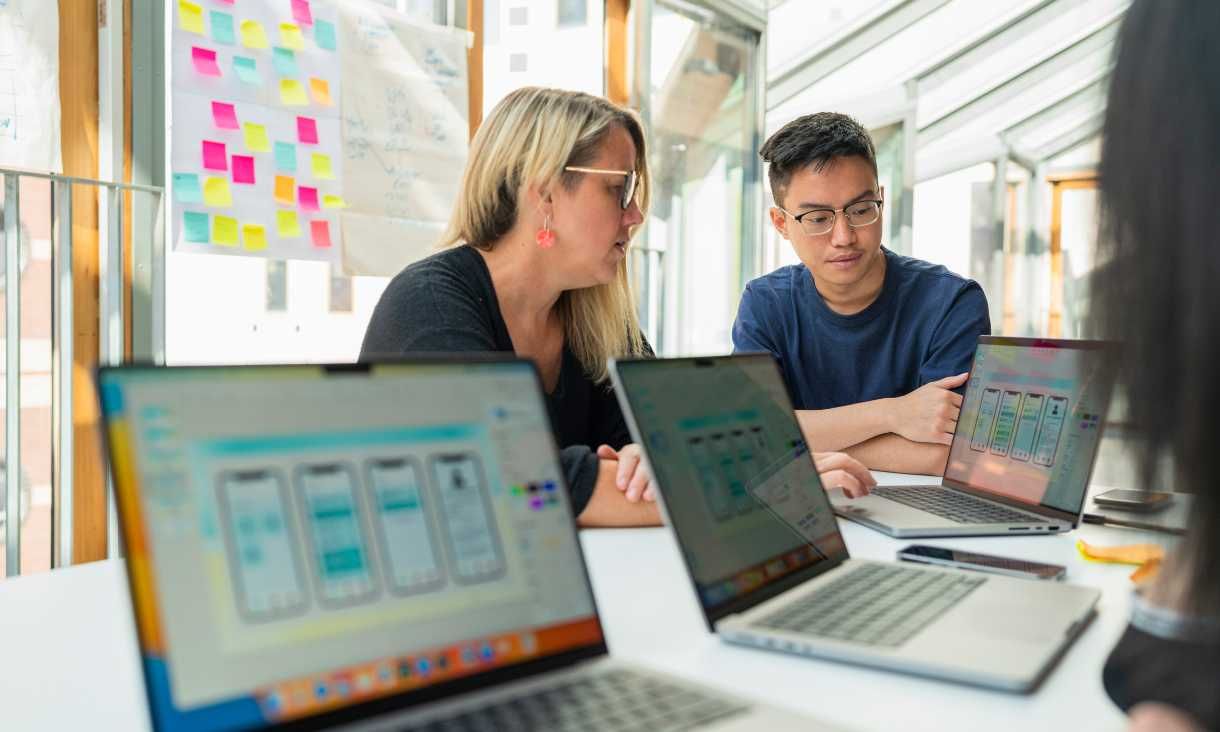 Woman and young male student sitting at desk with several laptops open in front of them learning the Apple Foundation Program.
